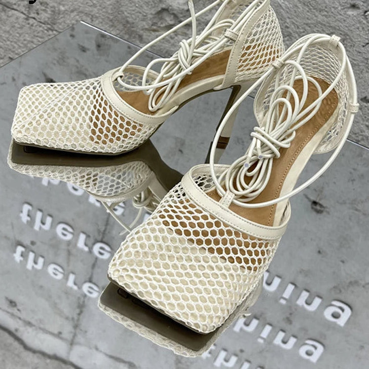 Beige high-heeled shoes with mesh design on a reflective surface.