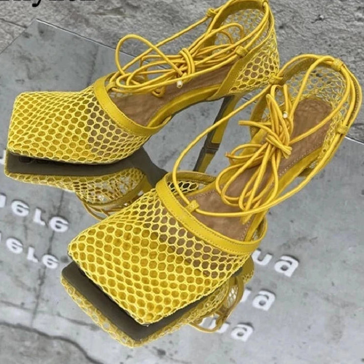 Yellow high-heeled shoes with mesh design on a reflective surface.