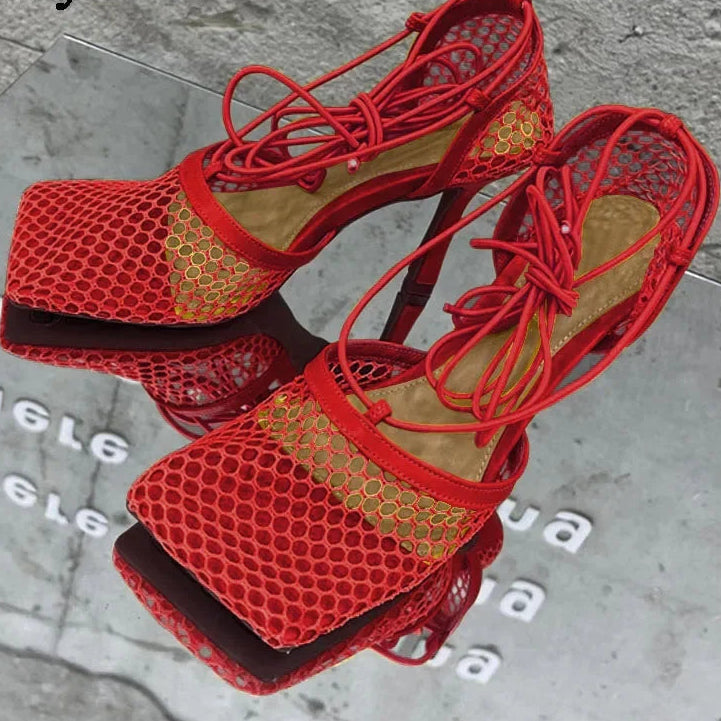 Red high-heeled sandals with mesh design on a reflective surface.
